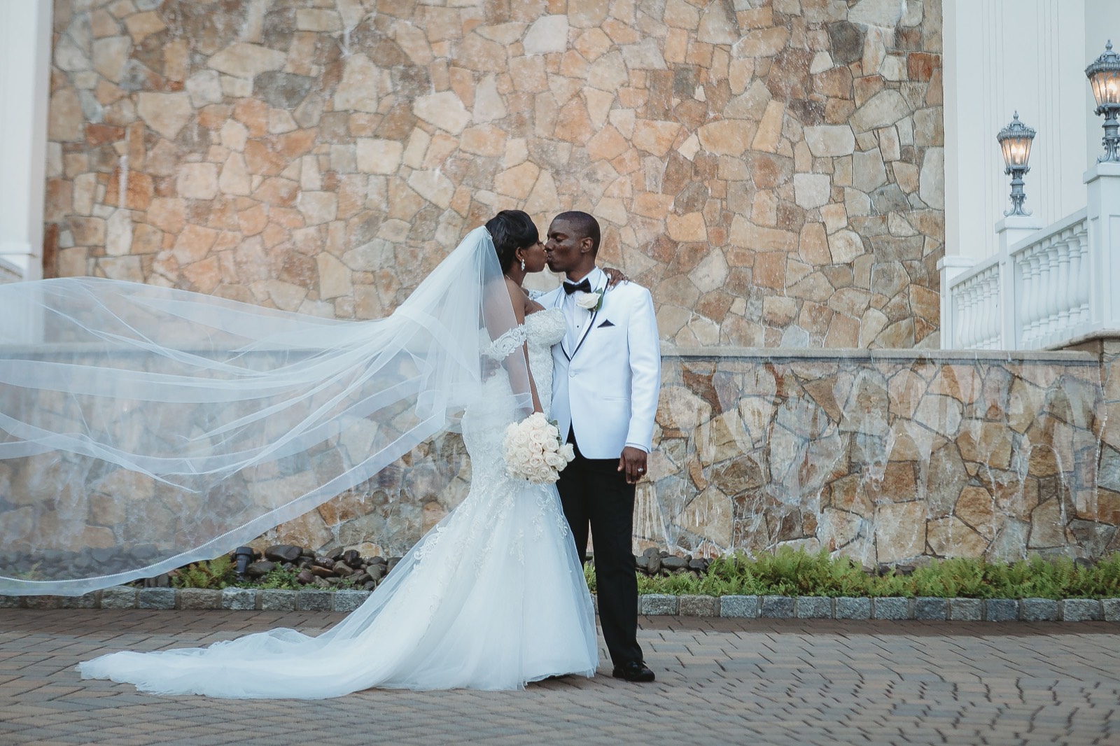 The bride and groom kissing by a stone wall as her veil sweeps dramatically in the wind.