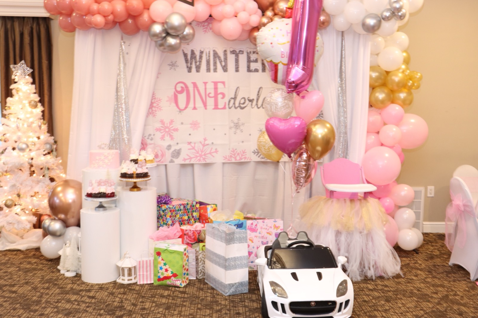 A pink 'Winter ONEderland' first-birthday backdrop with snowflakes, a tiered cake, cupcakes, gifts, and a flocked tree.